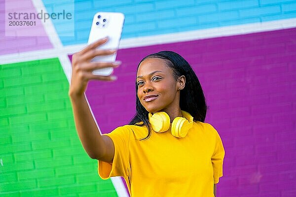 Niedliche afrikanische Frau  die ein Selfie mit einem Mobiltelefon macht  das neben einer bunten Wand steht