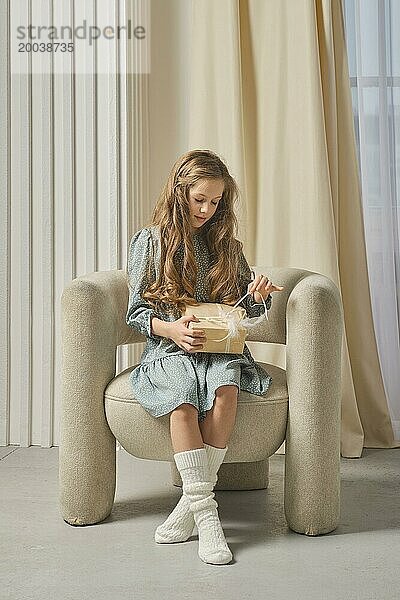 Ein junges Mädchen sitzt auf einem bequemen Stuhl und packt ein Geschenk in einem gut beleuchteten  gemütlichen Wohnzimmer aus. Die weichen Texturen und die warme Beleuchtung schaffen eine friedliche und heitere Atmosphäre