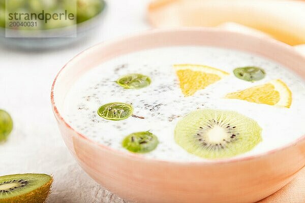 Joghurt mit Kiwi  Stachelbeere und Chia in einer Keramikschale auf grauem Betonhintergrund und orangefarbenem Leinentextil. Seitenansicht  Nahaufnahme  selektiver Fokus