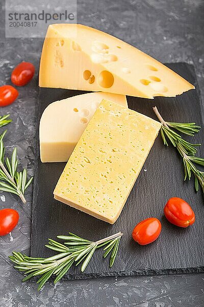 Verschiedene Käsesorten mit Rosmarin und Tomaten auf einer schwarzen Schiefertafel auf einem schwarzen Betonhintergrund. Seitenansicht  Nahaufnahme  selektiver Fokus