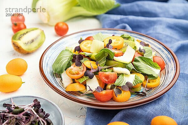 Vegetarischer Salat aus Pac Choi Kohl  Kiwi  Tomaten  Kumquat  Mikrogrünsprossen auf weißem Betonhintergrund und blauem Leinentuch. Seitenansicht  Nahaufnahme  selektiver Fokus