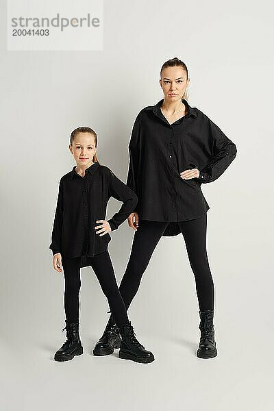 Eine moderne Familie in stilvollen schwarzen Kleidern posiert selbstbewusst vor einem schlichten weißen Hintergrund. Die Mutter und ihre Tochter tragen passende schwarze Stiefel  Hemden und enge Hosen