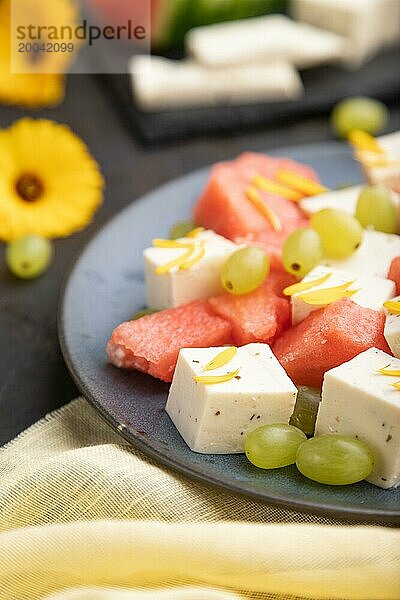 Vegetarischer Salat mit Wassermelone  Fetakäse und Trauben auf blauem Keramikteller auf schwarzem Betonhintergrund und gelbem Leinentuch. Seitenansicht  Nahaufnahme  selektiver Fokus