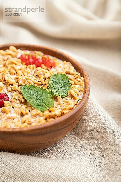 Weizenflockenbrei mit Milch  Himbeeren und Johannisbeeren in einer Holzschale auf weißem Holzhintergrund und Leinentextil. Seitenansicht  Nahaufnahme  selektiver Fokus