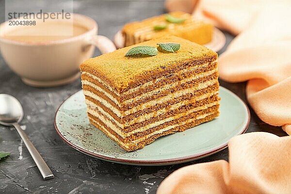 Hausgemachter Honigkuchen mit Milchcreme und Minze mit einer Tasse Kaffee auf einem schwarzen Betonhintergrund und orangefarbenem Textil. Seitenansicht  Nahaufnahme  selektiver Fokus