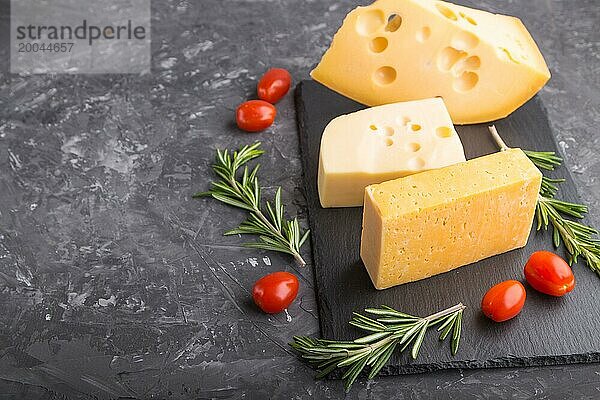 Verschiedene Käsesorten mit Rosmarin und Tomaten auf einer schwarzen Schiefertafel auf einem schwarzen Betonhintergrund. Seitenansicht  Nahaufnahme  Kopierraum  selektiver Fokus