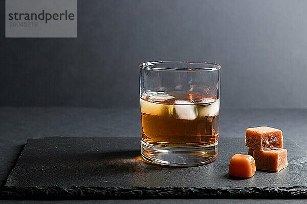 Glas bernsteinfarbener Whiskey mit Eis und Karamellbonbons auf einer schwarzen Schiefertafel aus Stein auf schwarzem Hintergrund. Seitenansicht  Nahaufnahme  selektiver Fokus  Kopierraum