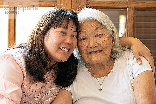 Nahaufnahme einer japanischen erwachsenen Tochter  die ihre grauhaarige Mutter lächelnd umarmt und in die Kamera schaut