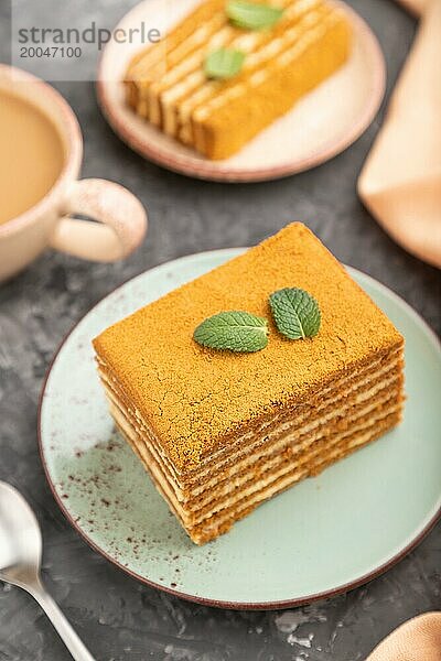 Hausgemachter Honigkuchen mit Milchcreme und Minze mit einer Tasse Kaffee auf einem schwarzen Betonhintergrund und orangefarbenem Textil. Seitenansicht  Nahaufnahme  selektiver Fokus