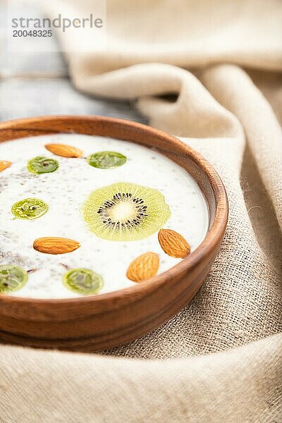 Joghurt mit Kiwi  Stachelbeere  Chia und Mandeln in einer Holzschale auf grauem Holzhintergrund und Leinenstoff. Seitenansicht  Nahaufnahme  selektiver Fokus