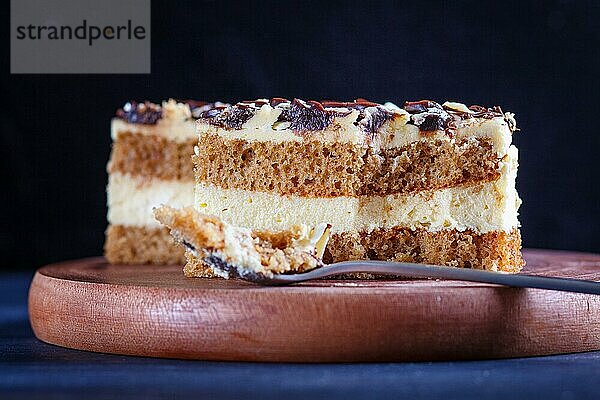 Ein Stück Kuchen mit Milch und Buttercreme  geschnitten mit einem Löffel auf einem hölzernen Küchenbrett  Nahaufnahme  selektiver Fokus  schwarzer Hintergrund