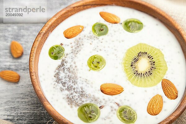 Joghurt mit Kiwi  Stachelbeere  Chia und Mandeln in einer Holzschale auf grauem Holzhintergrund und Leinenstoff. Seitenansicht  Nahaufnahme  selektiver Fokus