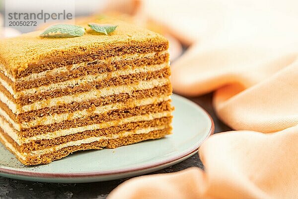 Hausgemachter Honigkuchen mit Milchcreme und Minze mit einer Tasse Kaffee auf einem schwarzen Betonhintergrund und orangefarbenem Textil. Seitenansicht  Nahaufnahme  selektiver Fokus