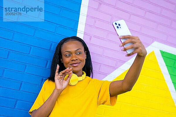 Lächelnde afrikanische Frau winkt  während sie ein Selfie mit dem Mobiltelefon vor einer bunten Wand macht