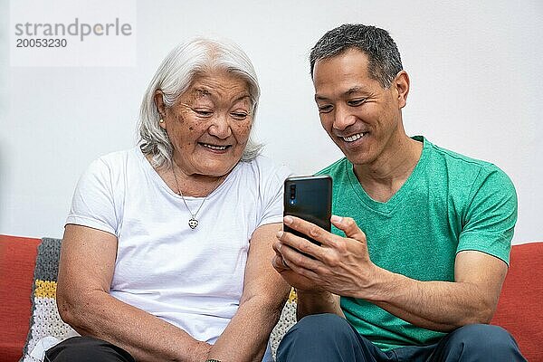 Erwachsener Sohn bringt seiner älteren Mutter japanischer Herkunft bei  wie man ein Mobiltelefon benutzt. Ältere Mutter und lateinisch japanischer Sohn schauen auf das Telefon und lächeln  genießen die gemeinsame Zeit zu Hause