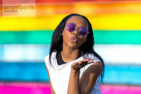 Afrikanische Frau mit Sonnenbrille  die gestikulierend einen Kuss in einem mehrfarbigen  lebhaften städtischen Hintergrund ausstößt