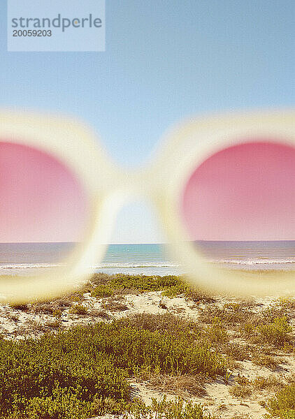 Ausgeschnittene Ansicht einer rosafarbenen Sonnenbrille mit Strand im Hintergrund