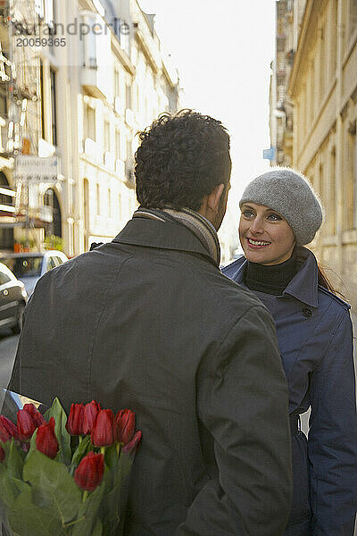 Rückansicht eines Mannes  der Blumen hinter seinem Rücken hält und vor einer Frau steht