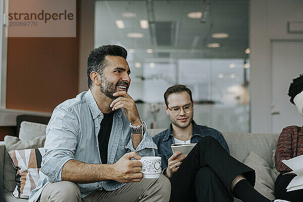 Lächelnder reifer Geschäftsmann mit Kaffeetasse  der während des Meetings neben einem männlichen Kollegen sitzt