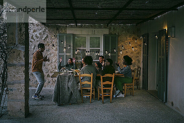 Männliche und weibliche Freunde sitzen am Esstisch und jubeln einem Mann zu  der während der Dinnerparty auf der Terrasse tanzt