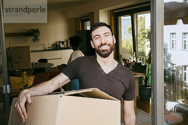 Porträt eines lächelnden Mannes  der einen Karton im neuen Zuhause trägt