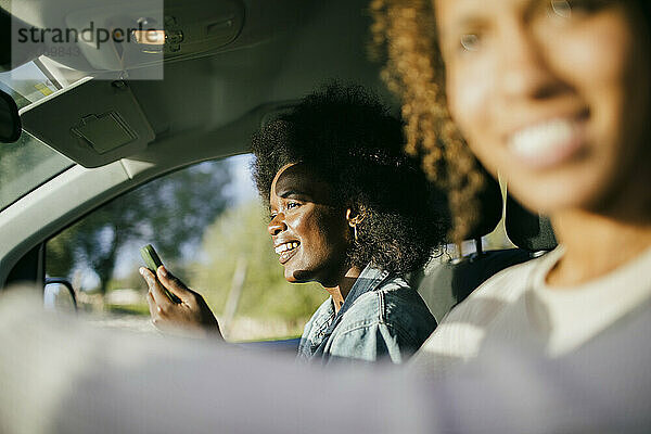 Lächelnde junge Frau mit Smartphone sitzt neben einem Freund im Auto