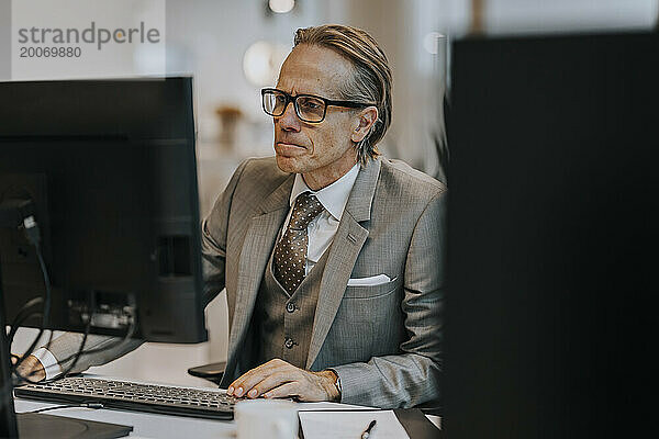 Reifer Geschäftsmann mit Brille und Computer am Schreibtisch im Büro