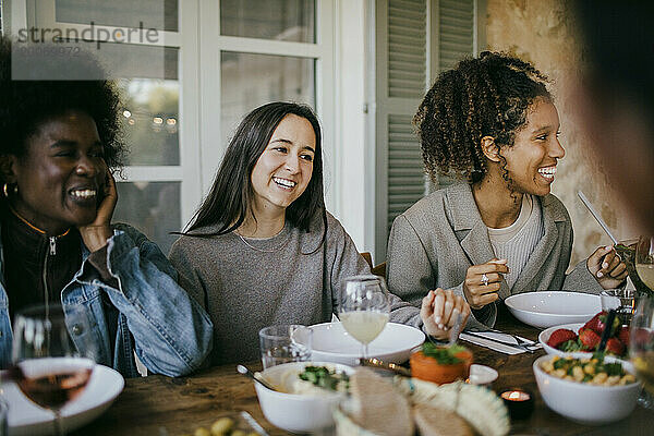 Fröhliche Freundinnen sitzen zusammen am Esstisch und genießen das Abendessen auf der Terrasse