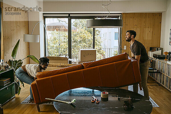 Gemischtrassiges Paar hilft einander beim Heben des Sofas im Wohnzimmer des neuen Zuhauses