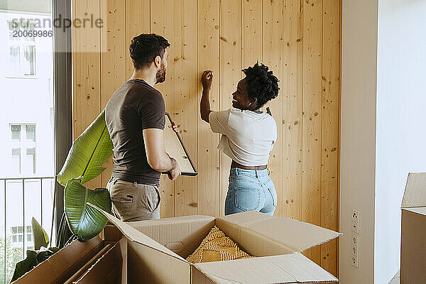 Glückliche Frau hämmert Nägel mit Freund  der Rahmen in der Nähe von Holzwand im neuen Zuhause hält