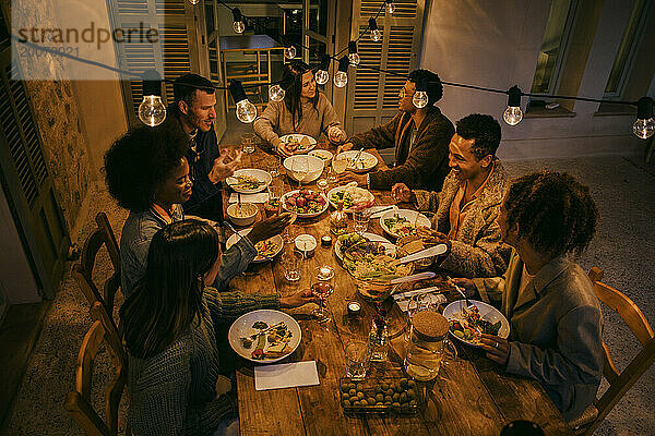 Blick aus der Vogelperspektive auf lächelnde Freunde  die während der Dinnerparty am beleuchteten Esstisch sitzen