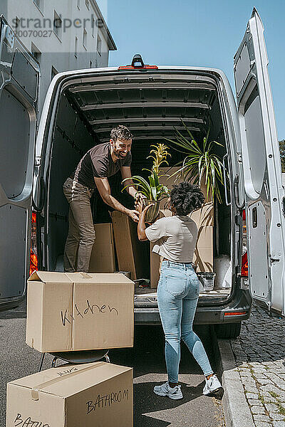 Gemischtrassiges Paar hilft sich gegenseitig beim Ausladen von Pflanzen aus dem Kofferraum eines Lieferwagens