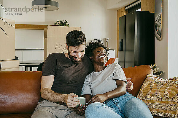 Glückliches gemischtrassiges Paar genießt es  sein Smartphone zu teilen  während es zu Hause auf dem Sofa im Wohnzimmer sitzt