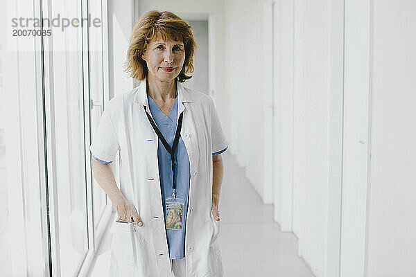 Porträt einer leitenden Ärztin  die mit den Händen in den Taschen auf dem Krankenhausflur steht