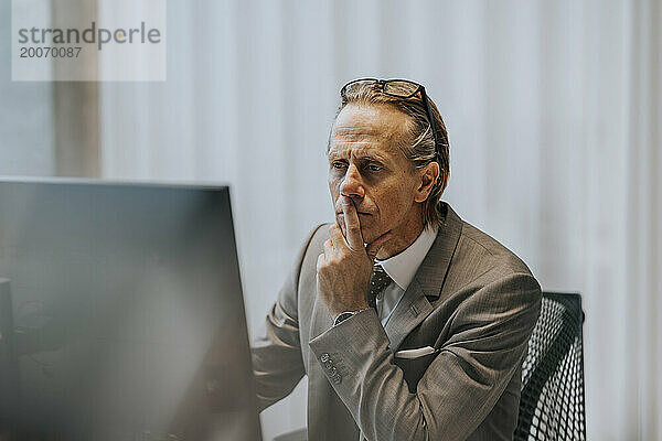 Reifer Geschäftsmann mit der Hand am Kinn und Blick auf den Computermonitor im Büro