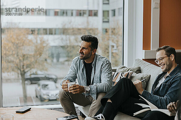 Lächelnder reifer Geschäftsmann mit Kaffeetasse  der während der Konferenzsitzung neben einem männlichen Kollegen sitzt