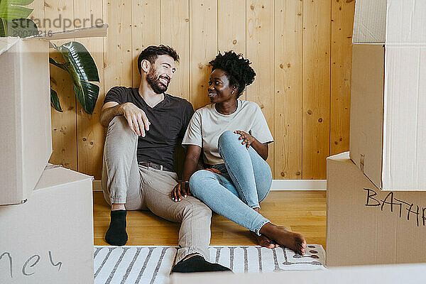 Glückliches gemischtrassiges Paar  das sich unterhält  während es zusammen in der Nähe einer Holzwand im neuen Zuhause sitzt