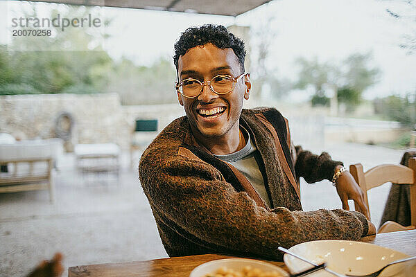 Glücklicher junger Mann mit Brille sitzt am Esstisch auf der Terrasse