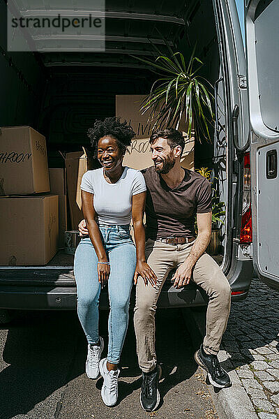 Glückliches gemischtrassiges Paar  das zusammen im Kofferraum eines Lieferwagens voller Kisten sitzt