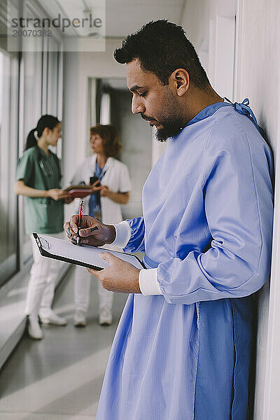 Seitenansicht eines männlichen Chirurgen  der im Krankenhausflur auf ein Klemmbrett schreibt