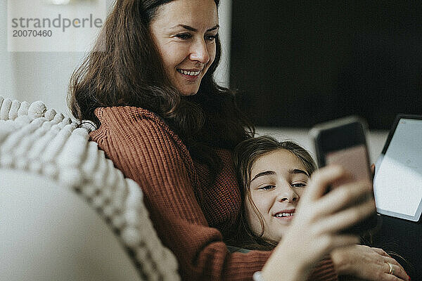 Mädchen teilt Smartphone mit lächelnder Mutter  während sie auf dem Sofa im Wohnzimmer liegt