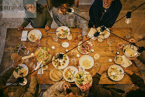 Blick aus der Vogelperspektive auf gemischtrassige Freunde  die während der Dinnerparty am beleuchteten Esstisch sitzen