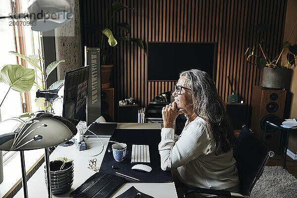 Seitenansicht einer lächelnden Geschäftsfrau mit der Hand am Kinn  die im Heimbüro einen Videoanruf führt