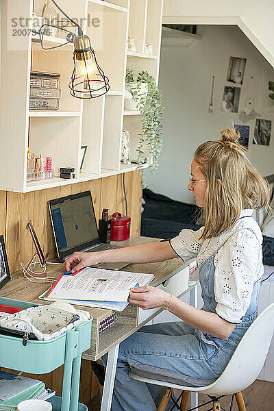 Blonde attraktive junge Frau sitzt an ihrem Schreibtisch in ihrem Zimmer und studiert auf ihrem Laptop