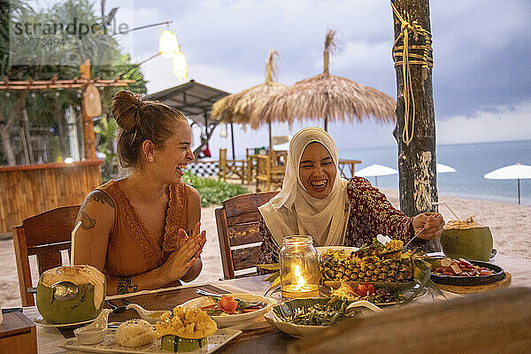 Zwei junge Freundinnen  eine trägt einen Hijab und isst in einem Strandrestaurant zu Abend  lacht und hat Spaß.