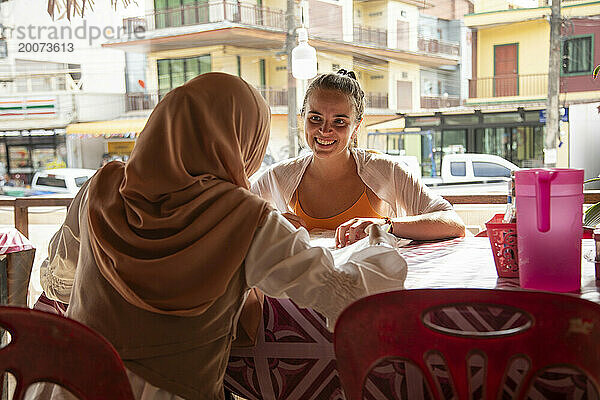 Zwei junge Frauen  eine davon trägt einen Hijab  sitzen zusammen in einem Café-Restaurant in einer ländlichen Stadt in Thailand