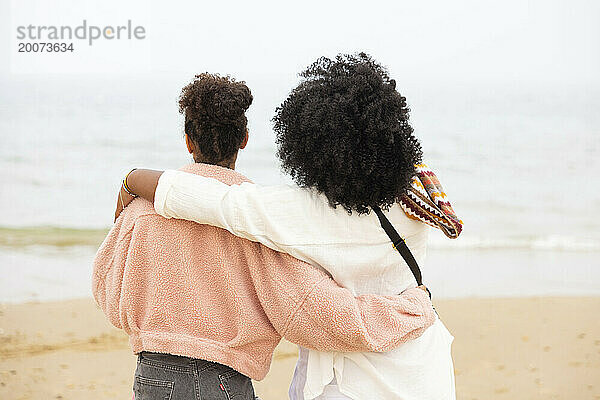Zwei gemischtrassige Schwestern am Strand umarmen sich und blicken auf das Meer.