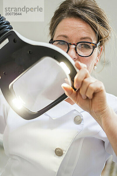 Porträt einer brünetten Kosmetikerin  die ihre Patientin durch eine Lupe betrachtet