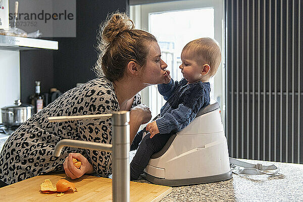 Mutter füttert ihr kleines Baby mit seinem Mittagessen  während es in seinem Hochstuhl oben auf der Küchenseite sitzt