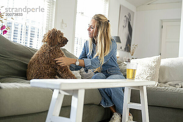 Attraktive blonde Frau sitzt auf ihrem Sofa und spricht mit ihrem Hund  der ihm Zuneigung schenkt